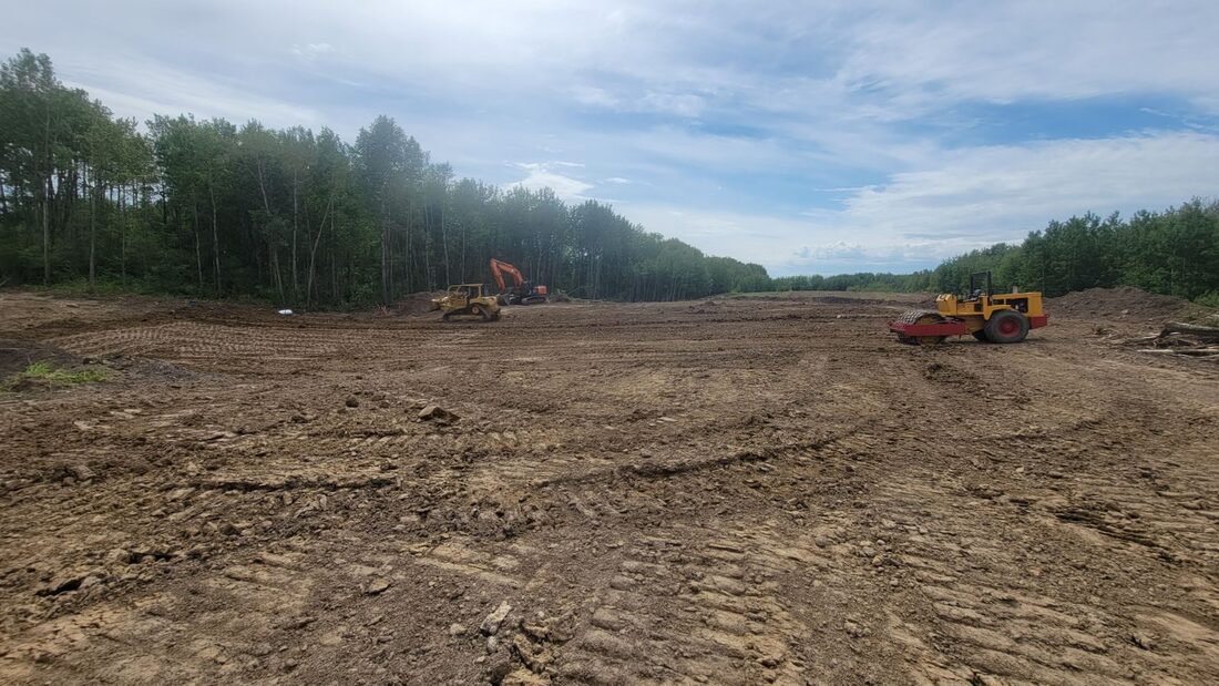 Cleared earthworks site with a packer in the foreground and a dozer and excavator working at the tree line